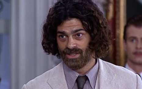 Eduardo Moscovis caracterizado como Petruchio. Ele têm o cabelo e barba longos, e as sobrancelha por fazer. O ator usa um terno branco e uma camisa azul em cena de O Cravo e a Rosa