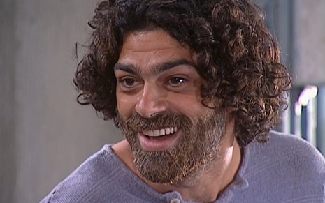 Eduardo Moscovis caracterizado como Petruchio: cabelo e barba longos, sobrancelha por fazer e blusa surrada. Ele sorri por conquista em cena de O Cravo e a Rosa.