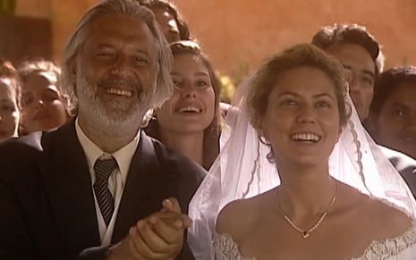 Patricia Pillar usa um vestido de noiva, e Antonio Fagundes, um terno; eles estão sorridentes em cena de casamento de O Rei do Gado