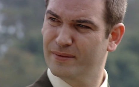 Jairo Mattos, caracterizado como o advogado Fausto, usa um terno cinza claro e dá um sorriso irônico em cena de O Rei do Gado