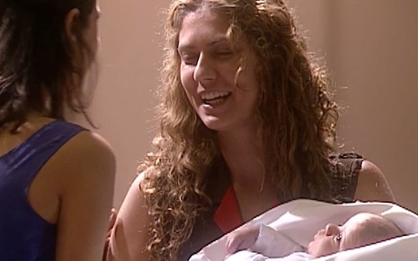 Patricia Pillar usa um vestido preto por cima de uma camiseta branca; ela parece tranquila enquanto segura um bebê nos braços em cena de O Rei do Gado