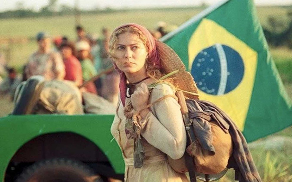 A atriz Patrícia Pillar com uma mochila nas costas, um lenço vermelho na cabeça, com um trator ao fundo com uma bandeira do Brasil, como Luana em O Rei do Gado