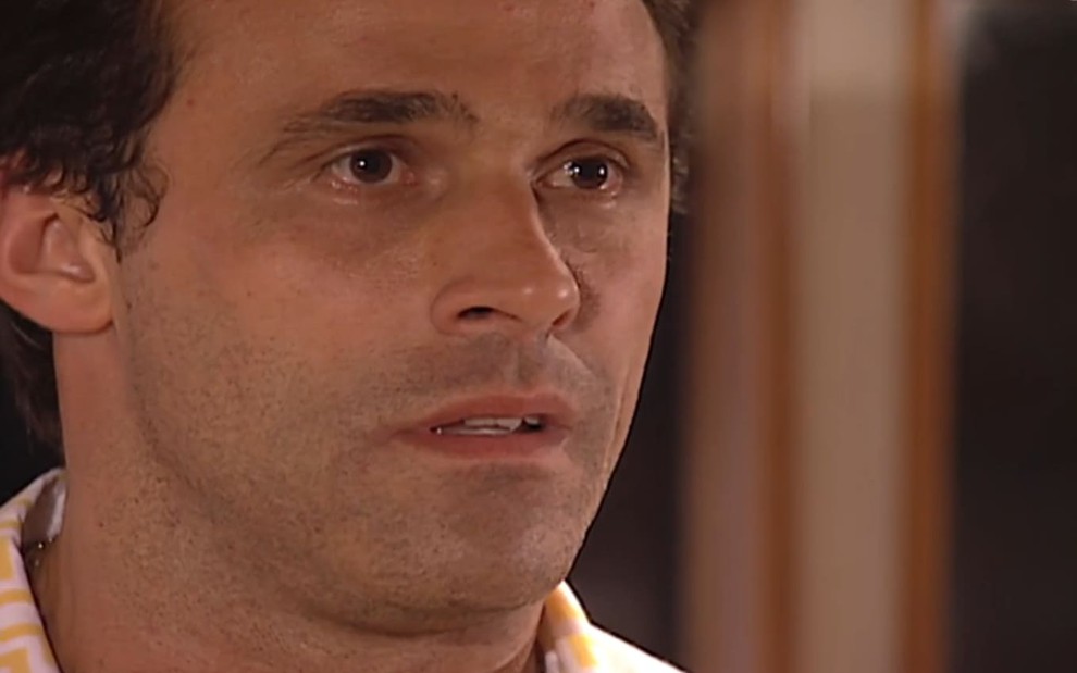 Oscar Magrini caracterizado como Ralf; ele usa uma camiseta roxa e dá um leve sorriso em cena da novela