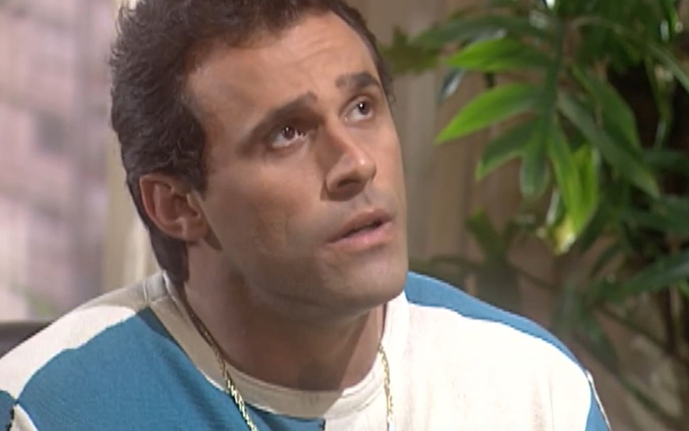 Oscar Magrini caracterizado como Ralf; ele usa uma camiseta branca e aparenta espanto em cena da novela