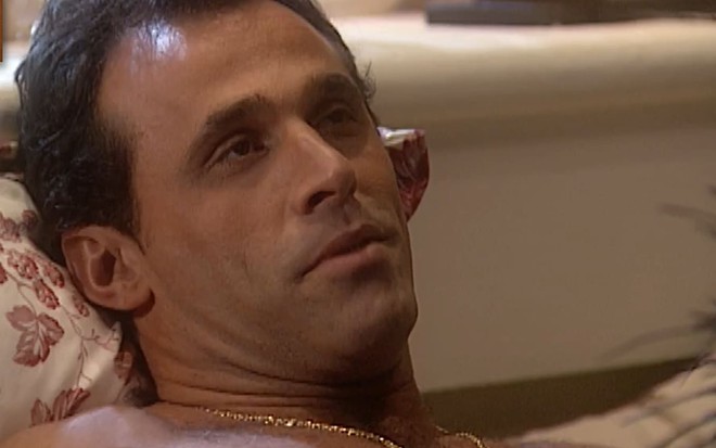 Oscar Magrini caracterizado como Ralf; ele usa uma camiseta branca e dá um leve sorriso em cena da novela