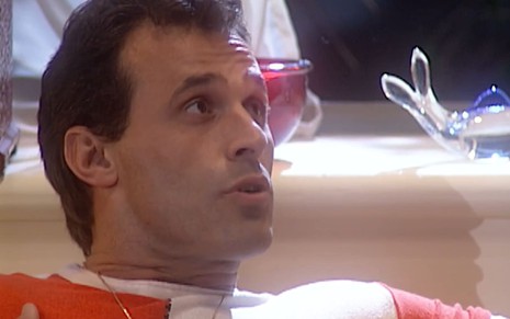 Oscar Magrini caracterizado como Ralf; ele usa uma camiseta branca e dá um leve sorriso em cena da novela