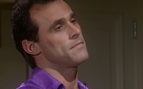 Oscar Magrini caracterizado como Ralf; ele usa uma camiseta roxa e dá um leve sorriso em cena da novela