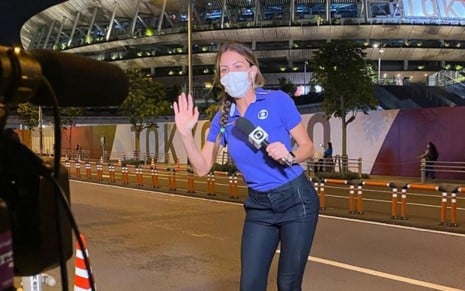 Bárbara Coelho está com o uniforme azul da Globo, fazendo um aceno de tchauzinho na frente da câmera