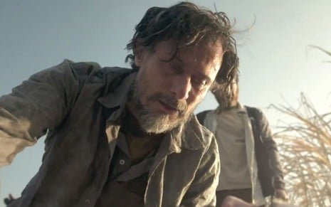 Enrique Diaz caracterizado como Gil em Pantanal: ator cabelo curto desalinhado, blusa de flanela cinza e se curva para alguém fora da tela