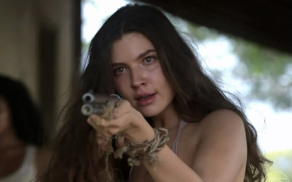 A atriz Alanis Guillen em cena de Pantanal na qual segura uma arma e faz uma expressão de braveza