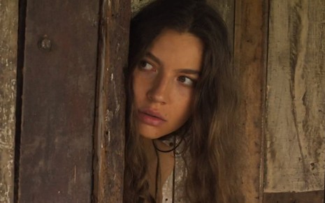 Juma (Alanis Guillen) está atrás de porta de tapera e observa escondida em cena de Pantanal, novela das nove da Globo