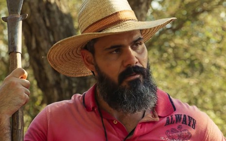 Juliano Cazarré, caracterizado como Alcides, tem a expressão séria em cena de Pantanal; ator segura uma zagaia e está posicionado em um campo, na frente de uma árvore