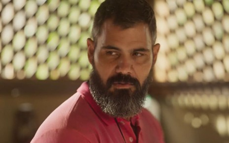 Juliano Cazarré, caracterizado como Alcides em Pantanal: ele tem a barba longa e grisalha. O ator veste uma camisa rosa, e o semblante está sério em cena de Pantanal