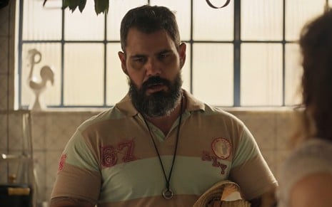 Juliano Cazarré, caracterizado como Alcides em Pantanal: ele tem a barba longa e grisalha. O ator veste um chapéu de palha e uma camisa polo rosa