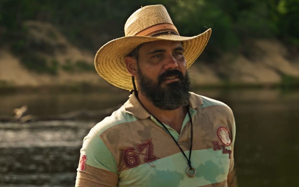 Juliano Cazarré, caracterizado como Alcides em Pantanal: ele tem a barba longa e grisalha. O ator está com chapéu de palha e uma camisa polo em cena de Pantanal