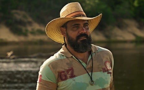 Juliano Cazarré, caracterizado como Alcides em Pantanal: ele tem a barba longa e grisalha. O ator está com chapéu de palha e uma camisa polo em cena de Pantanal