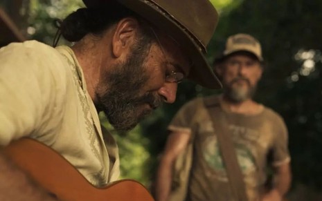 Almir Sater toca violão, caracterizado como Eugênio, em cena de Pantanal. Enrique Diaz o observa