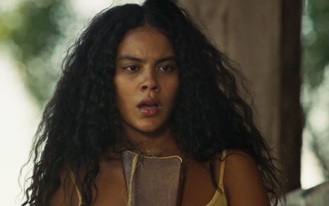 A atriz Bella Campos como Muda segura uma espingarda com expressão boquiaberta em cena de Pantanal