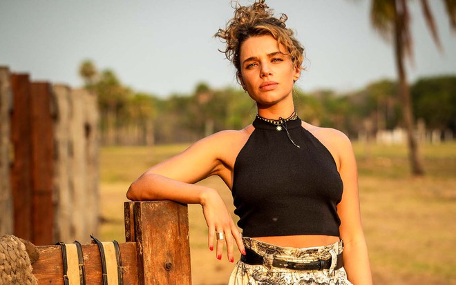A atriz Bruna Linzmeyer, como Madeleine, se apoia em uma porteira em um pasto em cena de Pantanal