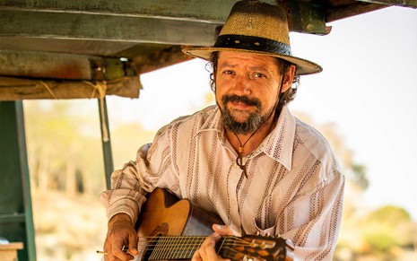 O ator Chico Teixeira em uma chalana com um violão no colo como o peão Quim em cena de Pantanal