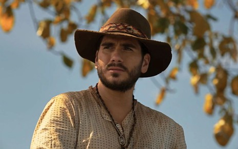 Gabriel Sater caracterizado como Trindade de Pantanal: ele usa um chapéu de palha e uma camisa surrada. Sério e com a barba bem aparada, ele tem o olhar distante em cena de Pantanal