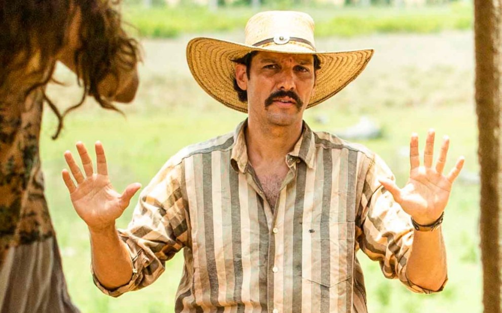 Guito, caracterizado como Tibério, veste camisa listrada e usa um chapéu de palha. O semblante está ameaçado, e as mãos para o alto em cena de Pantanal