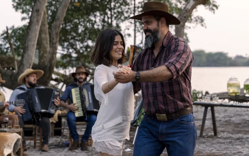 Juliano Cazarré, caracterizado como Alcides, usa uma camisa xadrez, uma calça jeans e um cinturão; ele está todo sorridente enquanto dança com Julia Dalavia, a Guta, em cena de Pantanal