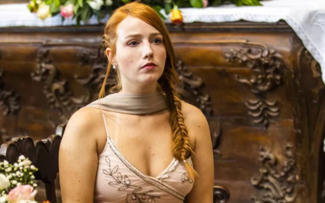 Malu Rodrigues, caracterizada como Irma, em cerimônia de casamento; ela tem o cabelo ruivo preso em uma trança lateral e usa um vestido bege decotado. Seu semblante está abatido em cena de Pantanal.