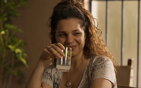 Isabel Teixeira, caracterizada como Maria Bruaca, exibe rosto sem maquiagem e cabelos amarrados; ela dá um sorriso sacana em cena de Pantanal. Com a mão direita, ela segura um copo de limonada quase vazio perto da boca