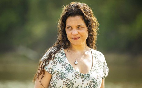 A atriz IsabelTeixeira faz um sorriso cínico em cena de Pantanal em que está com uma blusa estampada e os cabelos soltos como Maria Bruaca