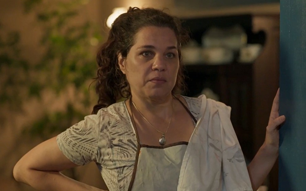 Isabel Teixeira, caracterizada como Maria Bruaca, exibe rosto sem maquiagem e cabelos amarrados; ela tem o semblante sério enquanto se apoia no batente da porta em cena de Pantanal