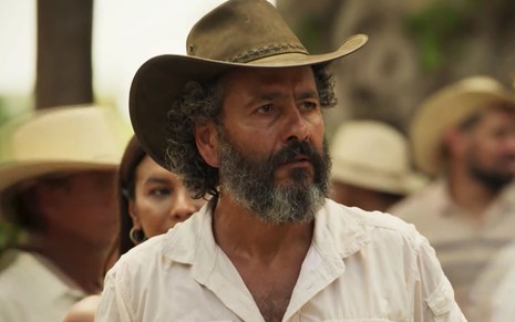 Marcos Palmeira em cena de Pantanal: ator está com camisa branca, chapéu de boiadeiro verde escuro e olha para alguém fora do quadro