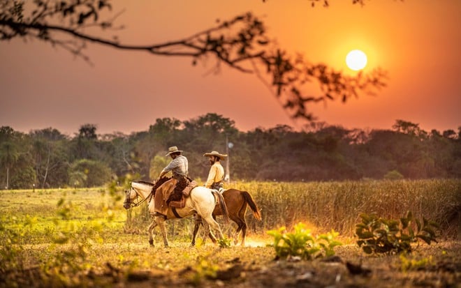 Renato Góes, caracterizado como José Leôncio, e Irandhir Santos, o Joventino, cavalgam em meio ao pôr do sol do Pantanal