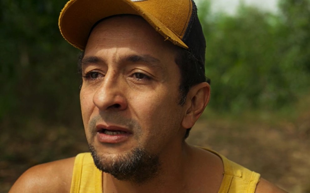 Irandhir Santos usa um boné marrom e uma regata amarela. Ele está emocionado em cena de Pantanal