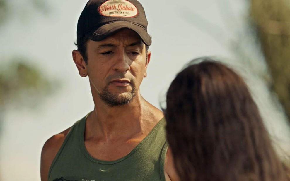 Irandhir Santos caracterizado como José Lucas: ele usa cavanhaque e veste uma regata verde. O cabelo cacheado está coberto por um boné marrom e bege em cena de Pantanal