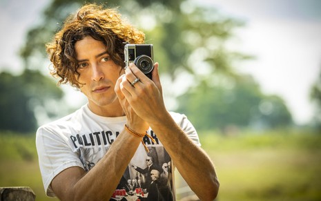 O ator Jesuíta Barbosa segura uma câmera fotográfica e olha para a frente com encantamento em cena da novela Pantanal