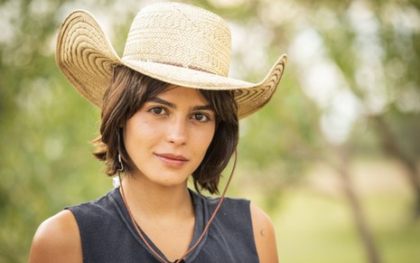 Julia Dalavia, caracterizada como Guta, encara a câmera com sorriso leve em ensaio fotográfico para Pantanal; ela veste regata e chapéu de palha