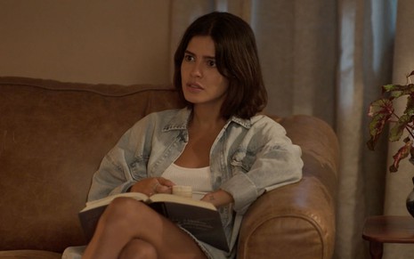 A atriz Julia Dalavia sentada em um sofá com um livro no colo e expressão de surpresa como a Guta em cena de Pantanal
