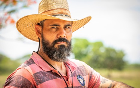 Juliano Cazarré, caracterizado como Alcides em Pantanal: ele tem a barba longa e grisalha. O ator veste um chapéu de palha e uma camisa polo rosa; o semblante está sério em ensaio fotográfico de divulgação de Pantanal
