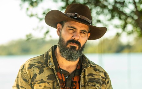 Juliano Cazarré usa um chapéu de couro, uma camisa preta e uma jaqueta de estampa militar; ele encara a câmera, sério, em ensaio de divulgação de Pantanal