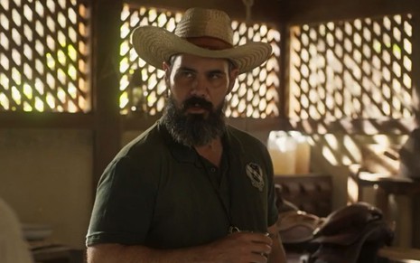 Alcides (Juliano Cazarré) está em pé e usa chapéu em cena de Pantanal