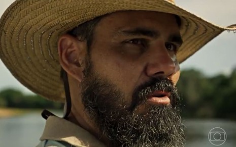 De chapéu, Alcides (Juliano Cazarré) está parado à beira do rio em cena de Pantanal, novela das nove da Globo