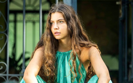 Alanis Guillen, caracterizada como Juma, tem a expressão pensativa; atriz está agachada em frente a porta da mansão Novaes em ensaio fotográfico de Pantanal