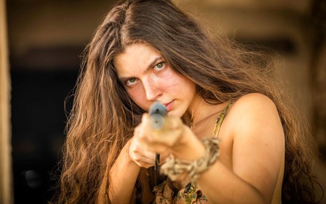 Alanis Guillen, caracterizada como Juma em Pantanal; atriz tem o rosto com manchas de sol, boca seca e cabelos castanhos soltos. Ela aponta a arma para a câmera em ensaio de divulgação de Pantanal