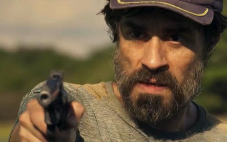 Erom Cordeiro em cena de Pantanal: ator está caracterizado como Lúcio e segura arma com a mão direita