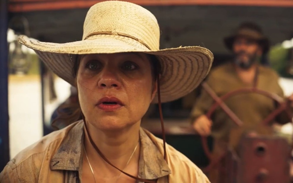 Isabel Teixeira usa um chapéu de palha e vestes surradas; ela tem a expressão chocada enquanto encara ao longe. É possível ver Almir Sater no fundo da imagem.