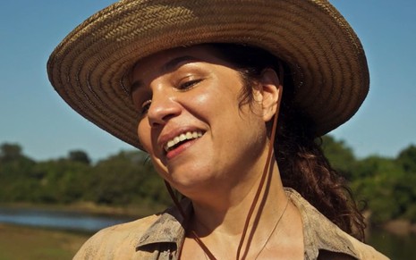Isabel Teixeira caracterizada como Maria Chalaneira: atriz usa um chapéu de palha e uma regata branca e tem os cabelos amarrados. Ela sorri, livre.
