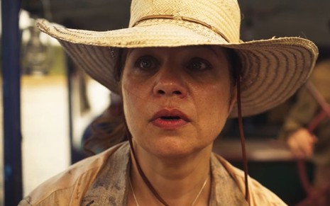 Isabel Teixeira, caracterizada como Maria Bruaca, usa camisa surrada e um chapéu de palha; ela tem o semblante chocado em cena de Pantanal
