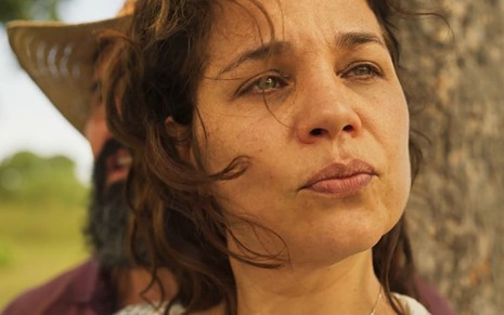 Isabel Teixeira caracterizada como Maria Bruaca: atriz não usa maquiagem e tem os cabelos presos. Ela encara um ponto fixo com o canto do olho e faz bico em cena de Pantanal