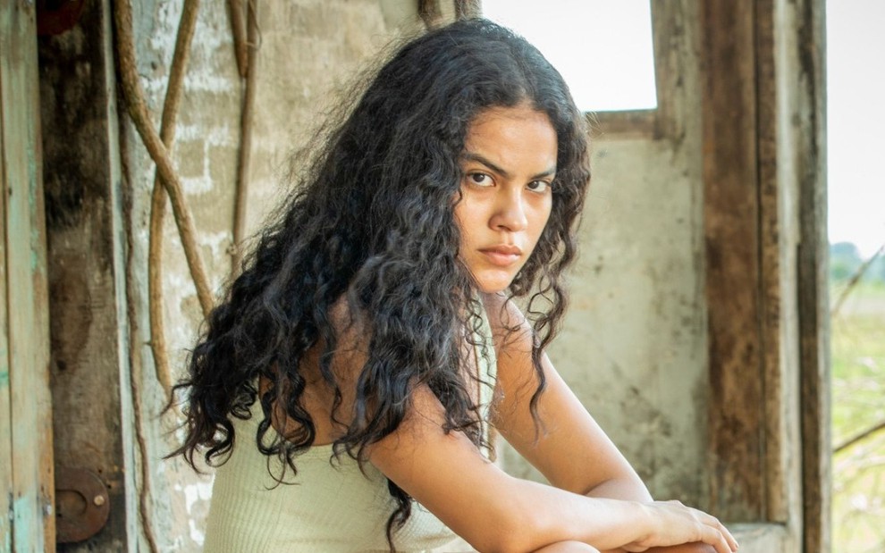 Bella Campos em cena de Pantanal: atriz está agachada, com os cabelos soltos, ela olha à câmera